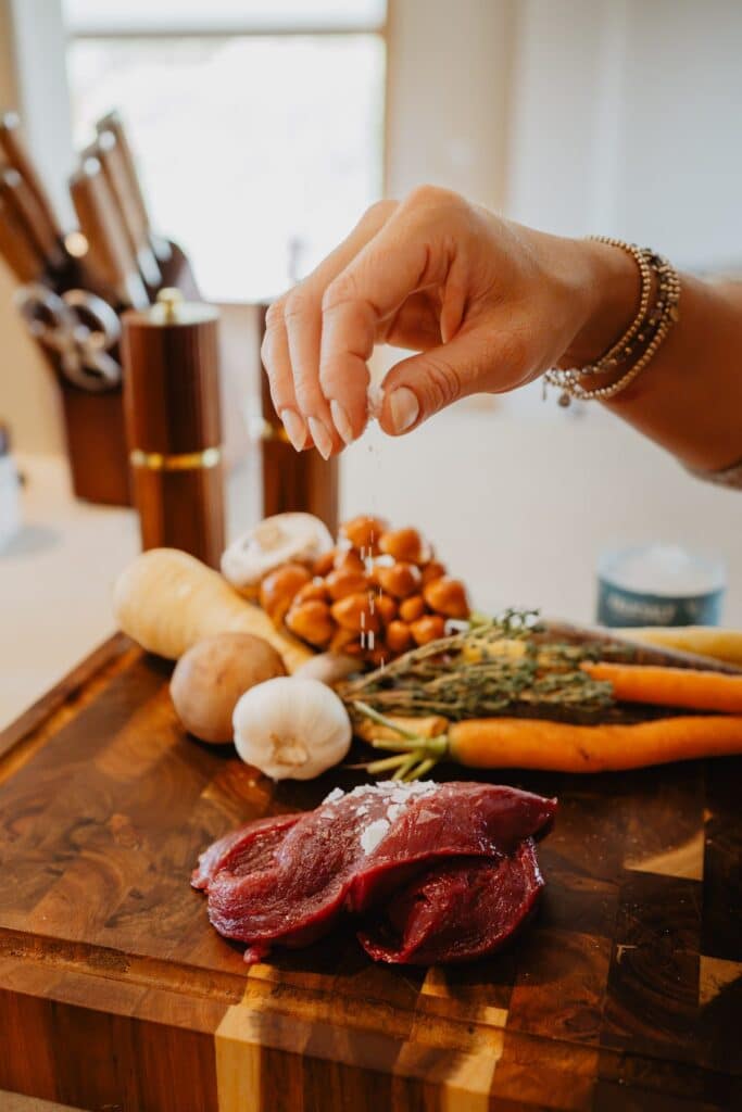 Hand strooit kruiden over wortels, pastinaak, knoflook en vlees op een houten snijplank